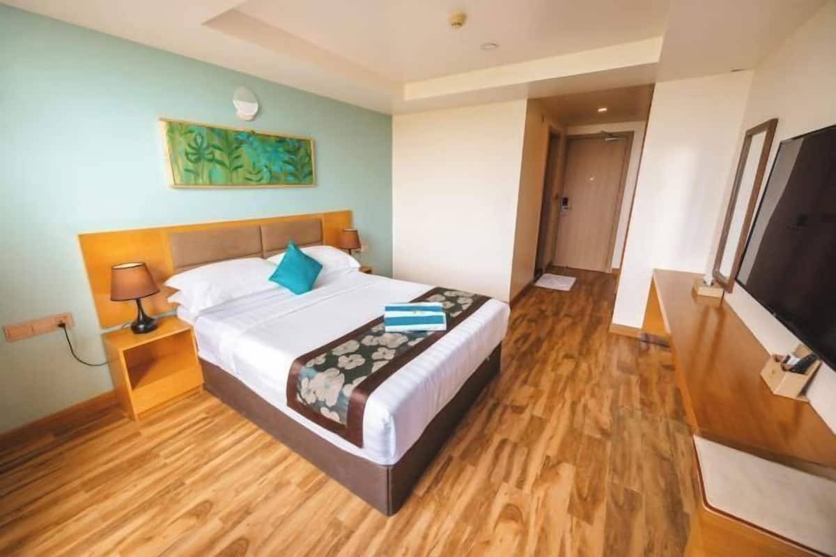 A hotel room with a wooden floor, a bed with white linens, a blue pillow, a floral throw blanket, a nightstand with a lamp, and a TV on a wooden stand.