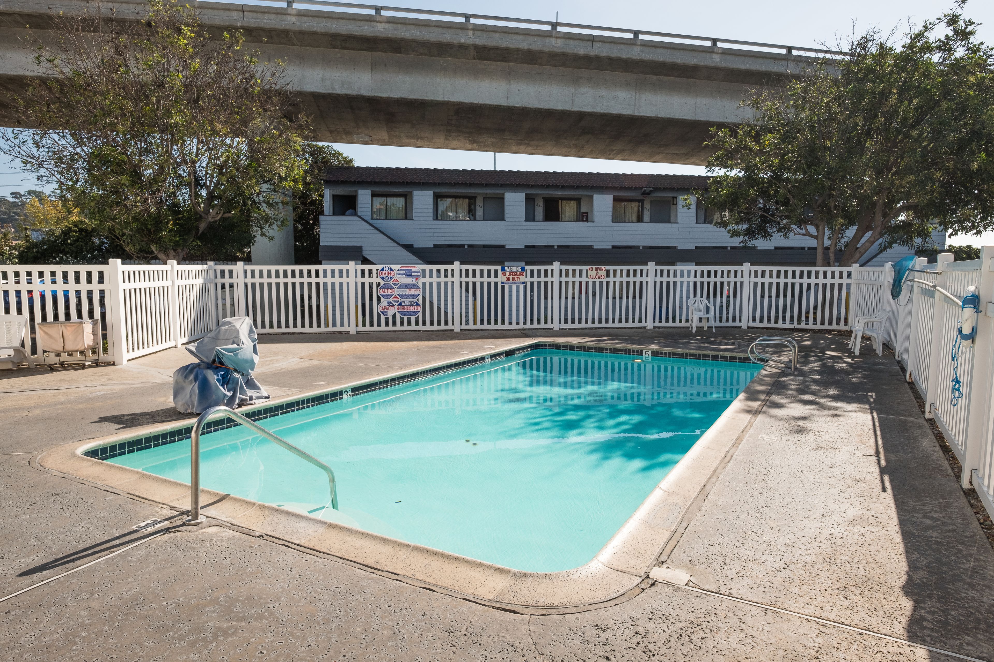 Una piscina rectangular con una escalera metálica, rodeada por una valla blanca, bajo un paso elevado.