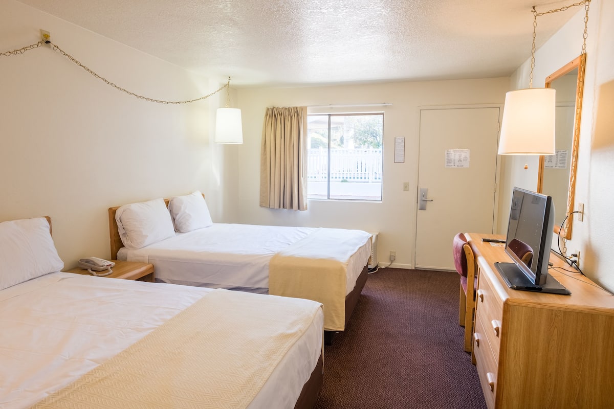 A hotel room with two beds, a desk with a computer, a lamp, and a window with curtains.