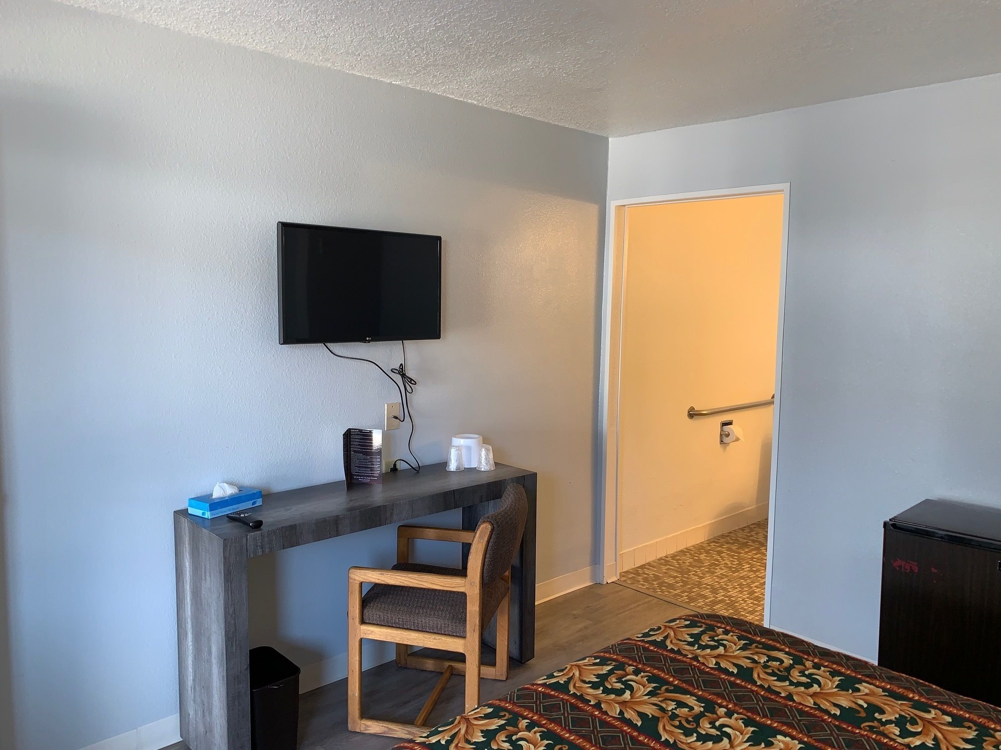 A hotel room with a wooden desk, a television mounted on the wall, a chair, a patterned rug, and a door.