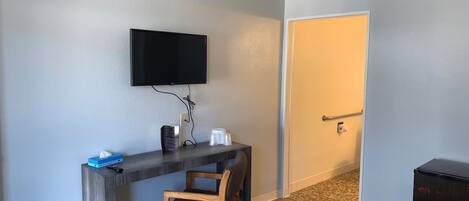 A hotel room with a wooden desk, a television mounted on the wall, a chair, a patterned rug, and a door.