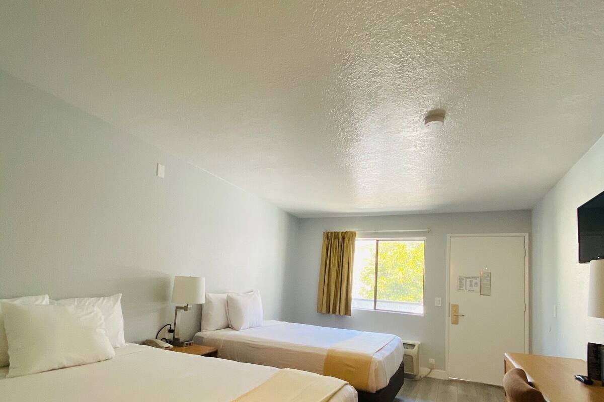 A hotel room with two beds, a desk, a chair, a window with curtains, and a door.