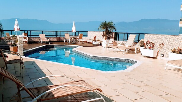A rooftop pool area with lounge chairs and umbrellas, overlooking a body of water and mountains. - Jangada Flat Service (Caraguatatuba)