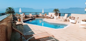 Un área de piscina en la azotea con sillones reclinables y sombrillas, con vistas a un cuerpo de agua y montañas.