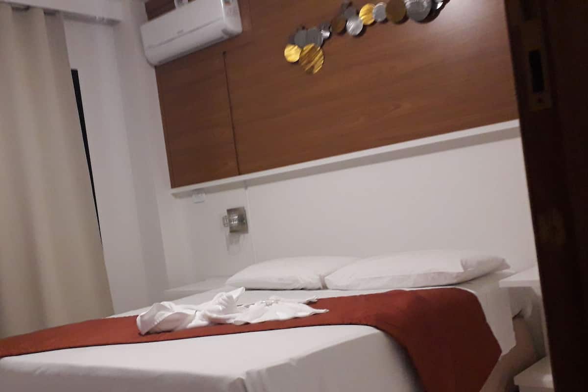 Habitación de hotel con cama, ropa de cama blanca, cabecero de madera y aire acondicionado instalado en la pared.
