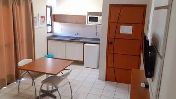 A compact kitchen with a microwave, sink, and a small refrigerator. There is a table with two chairs and a blue trash bin.