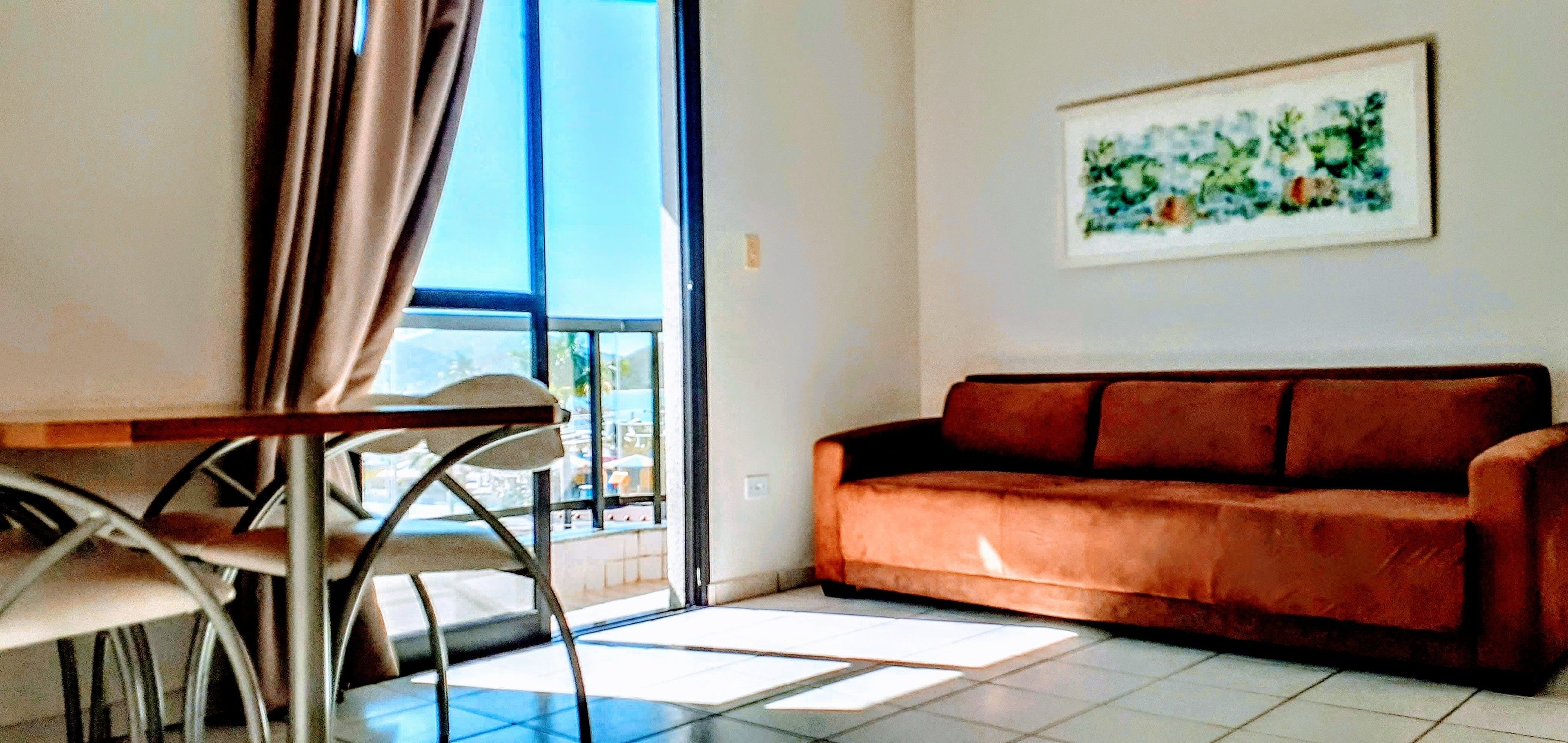 A living room with a brown sofa, a dining table with chairs, and a large window with curtains.