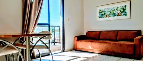 A living room with a brown sofa, a dining table with chairs, and a large window with curtains.