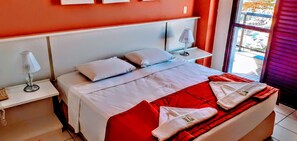 A hotel room with a bed, pillows, a nightstand, a lamp, and a window with a view. - Jangada Flat Service (Caraguatatuba)