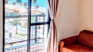 A room with a large window overlooking a street and beach, a brown sofa, and a painting on the wall.