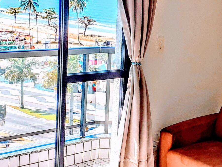 A room with a large window overlooking a street and beach, a brown sofa, and a painting on the wall.