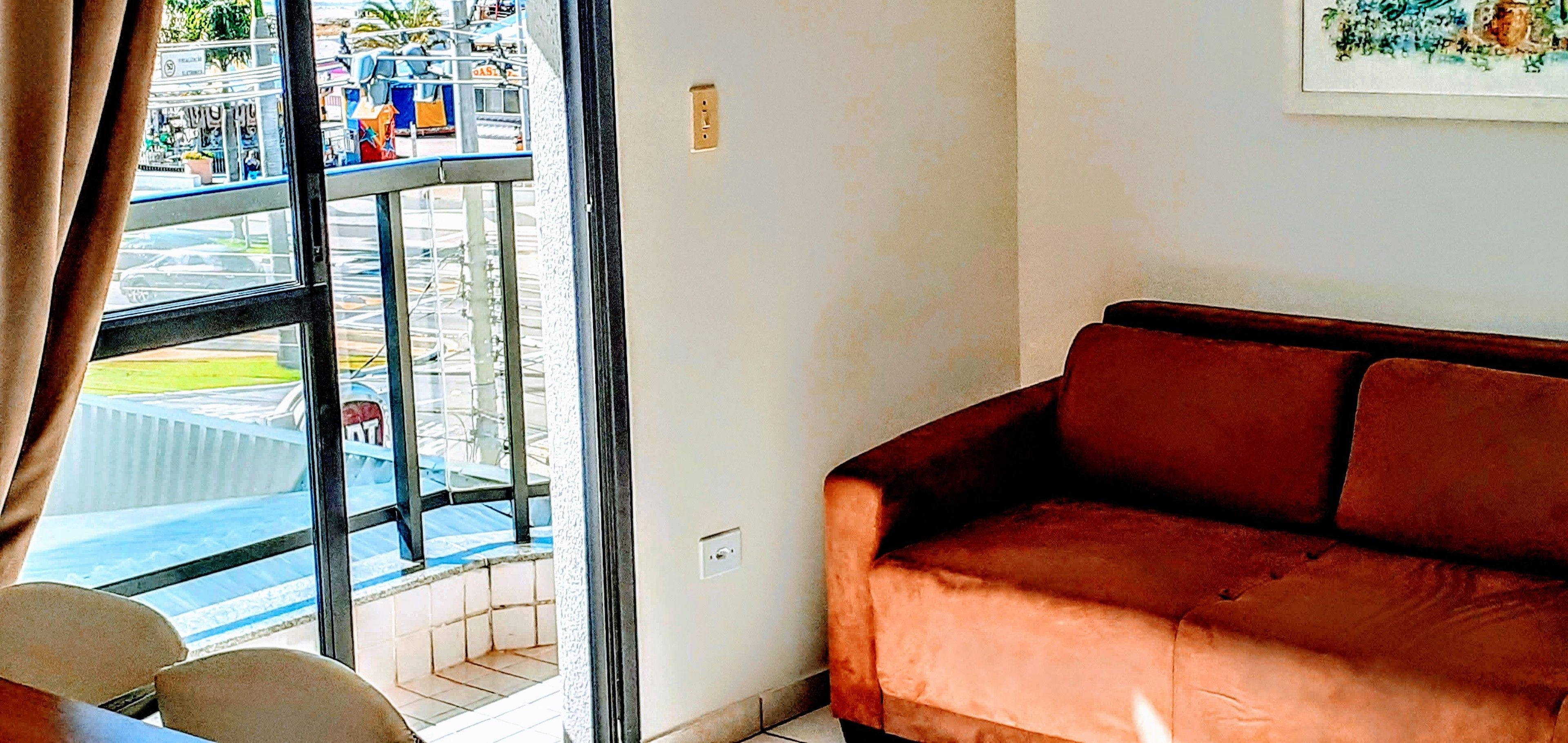 A living room with a brown sofa, a window with a view of a parking lot and a building, and a tiled floor.