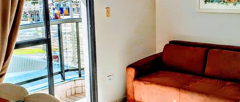 A living room with a brown sofa, a window with a view of a parking lot and a building, and a tiled floor.