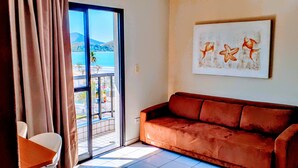 A room with a brown sofa, a painting of fish, a sliding glass door to a balcony, and a chair. - Jangada Flat Service (Caraguatatuba)
