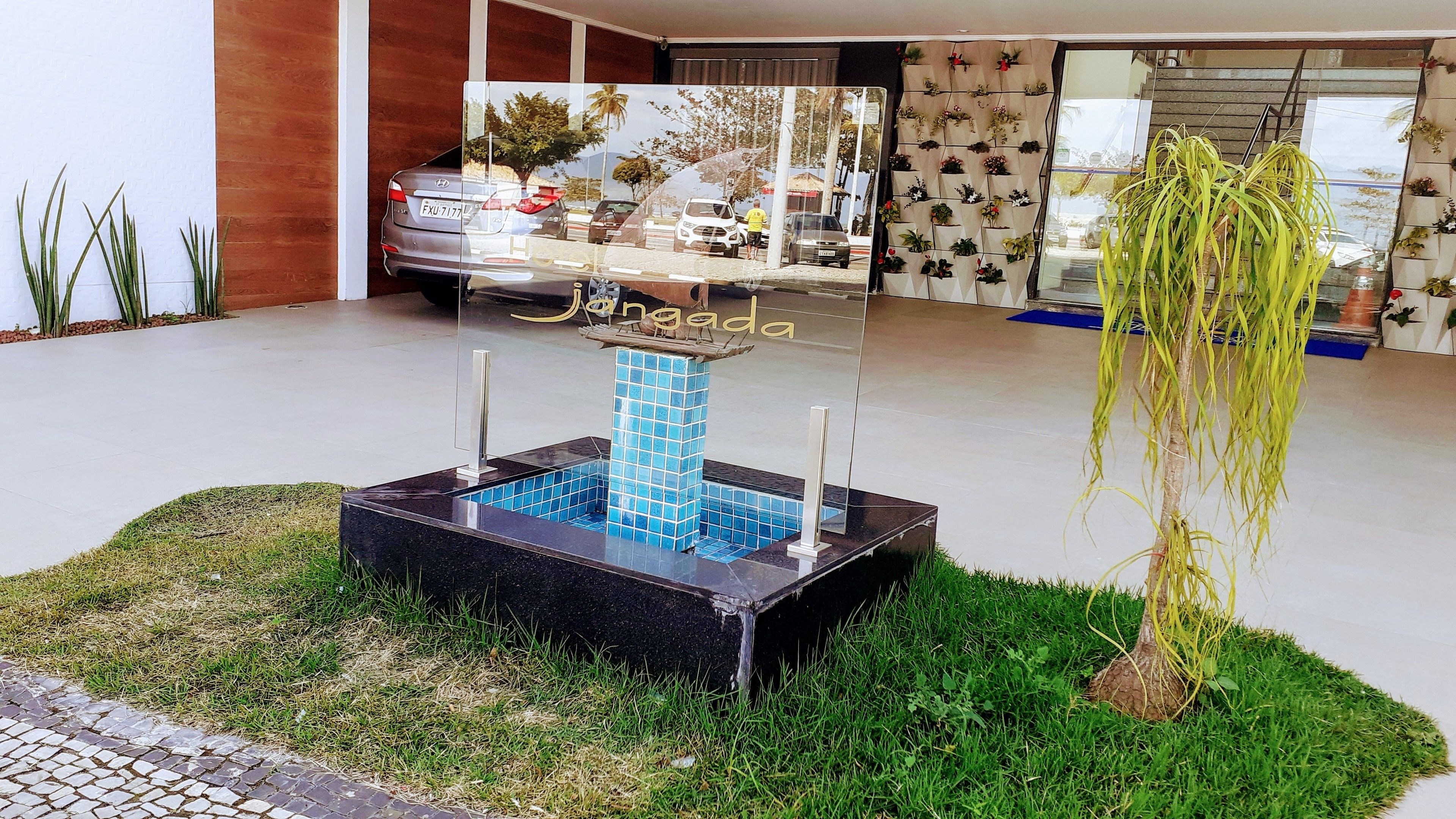 Un espacio exterior moderno con una fuente decorativa, un pequeño jardín y el reflejo de coches aparcados y la fachada de un edificio.