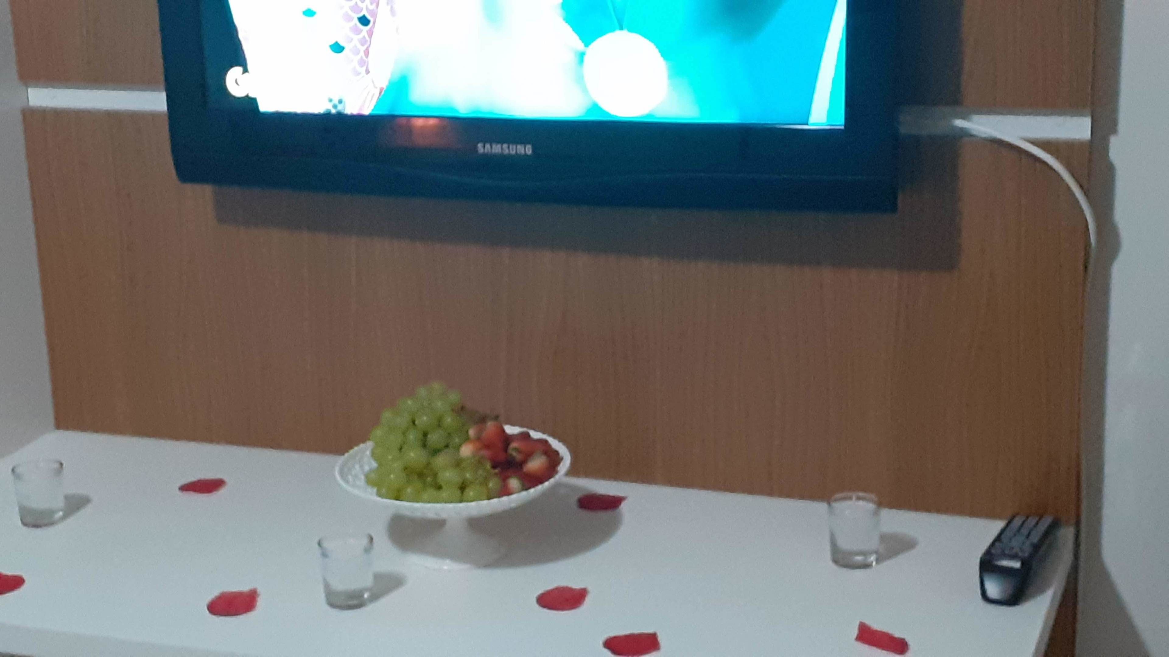 Egy falra szerelt Samsung televízió, egy asztal, rajta egy gyümölcstál, és egy távirányító.