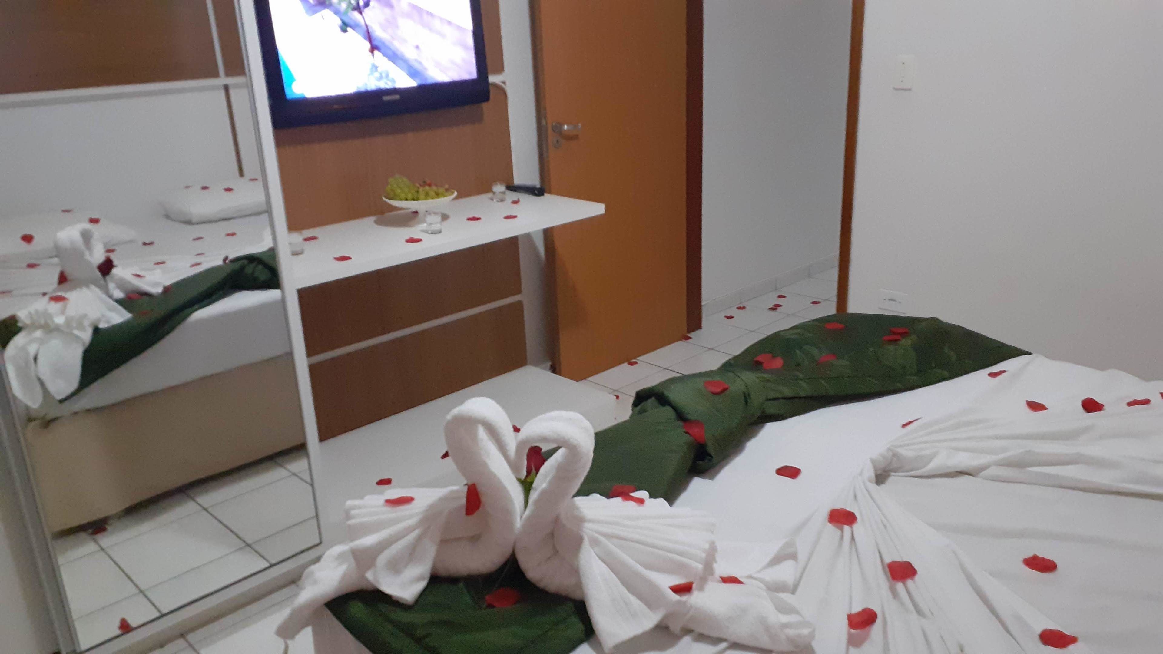 Quarto de hotel com cama, espelho, televisão e uma pequena mesa com uma tigela de frutas.