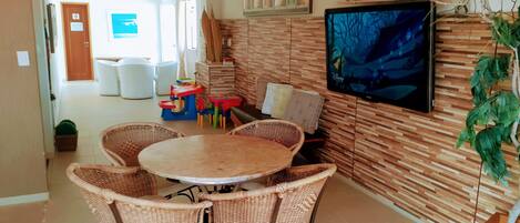 A dining area with wicker chairs and a round table, a television mounted on the wall, and a wooden wall with a patterned design.
