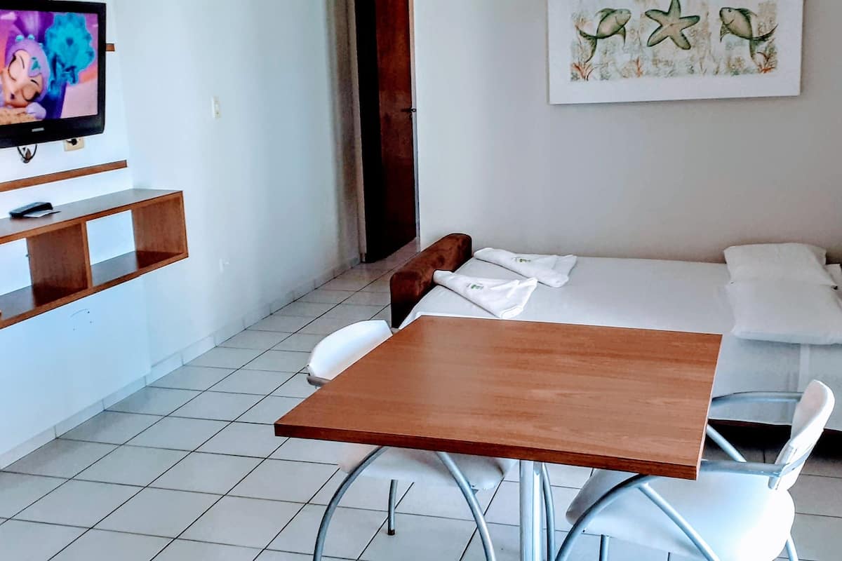 Una habitación pequeña y limpia, con una cama, una mesa y una silla. Hay un televisor en la pared y un cuadro sobre la cama.