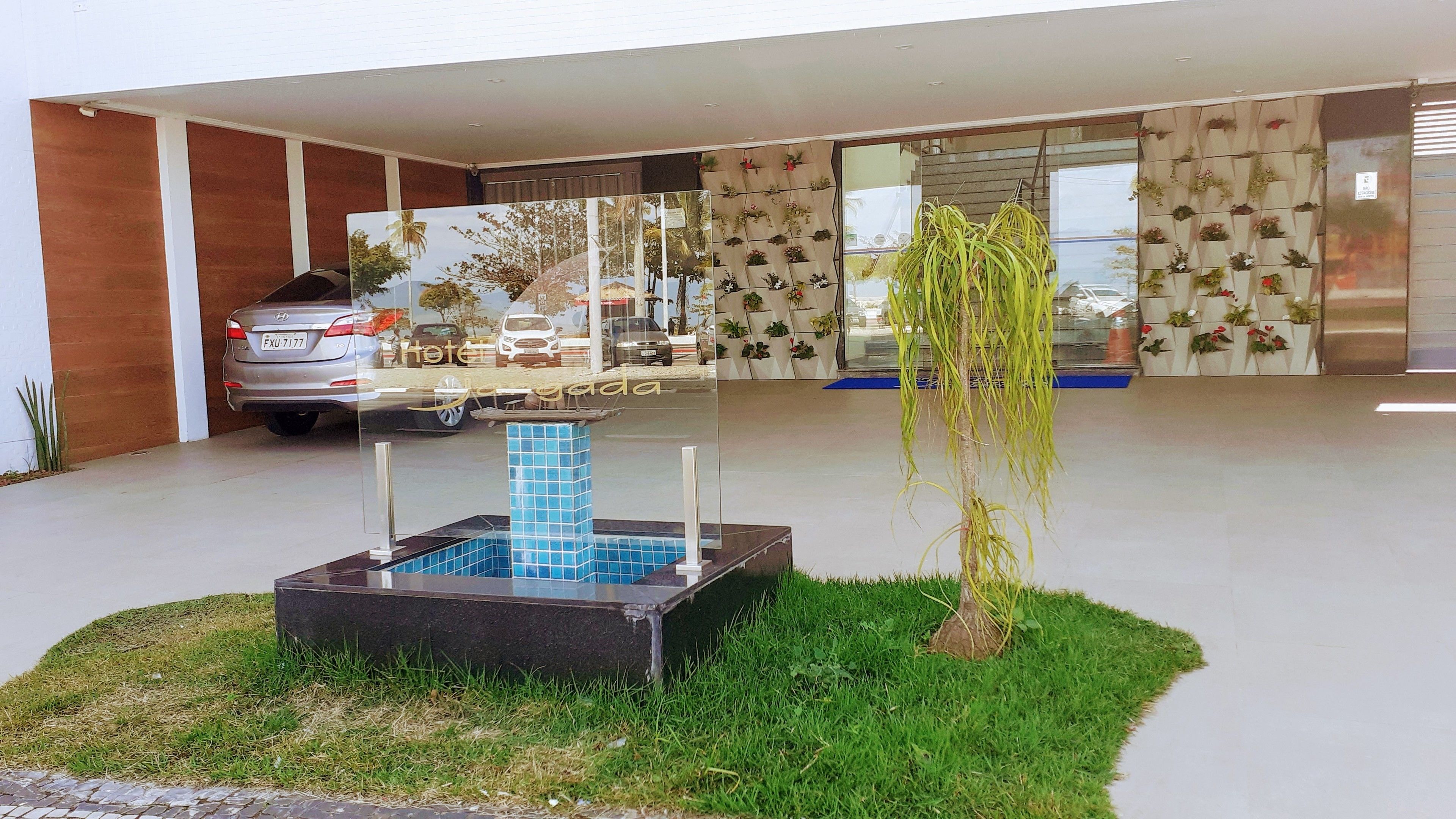 A modern hotel entrance with a reflective glass facade, a small outdoor water feature, and a parking area with a car.