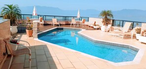 A rooftop pool with lounge chairs and umbrellas, surrounded by a brick wall and potted plants. - Jangada Flat Service (Caraguatatuba)