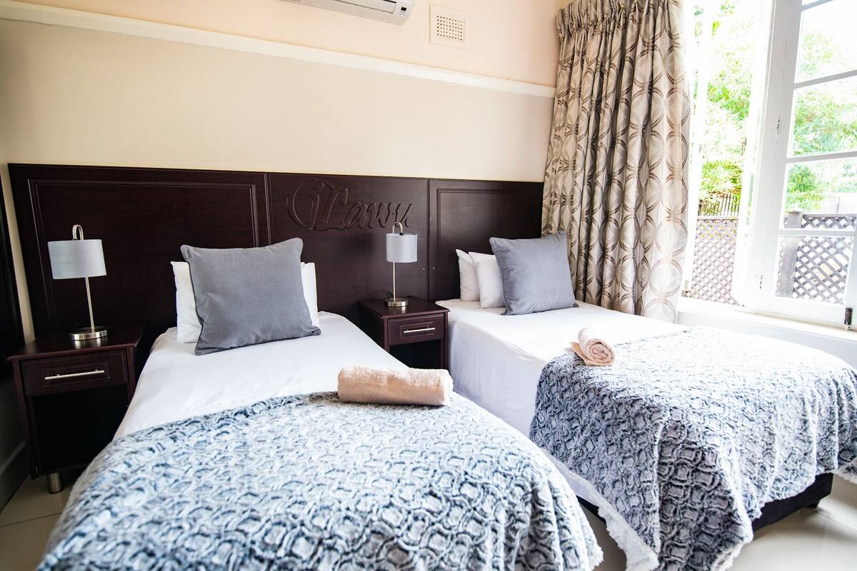 A hotel room with two beds, a dark wooden headboard, bedside lamps, and a window with curtains.