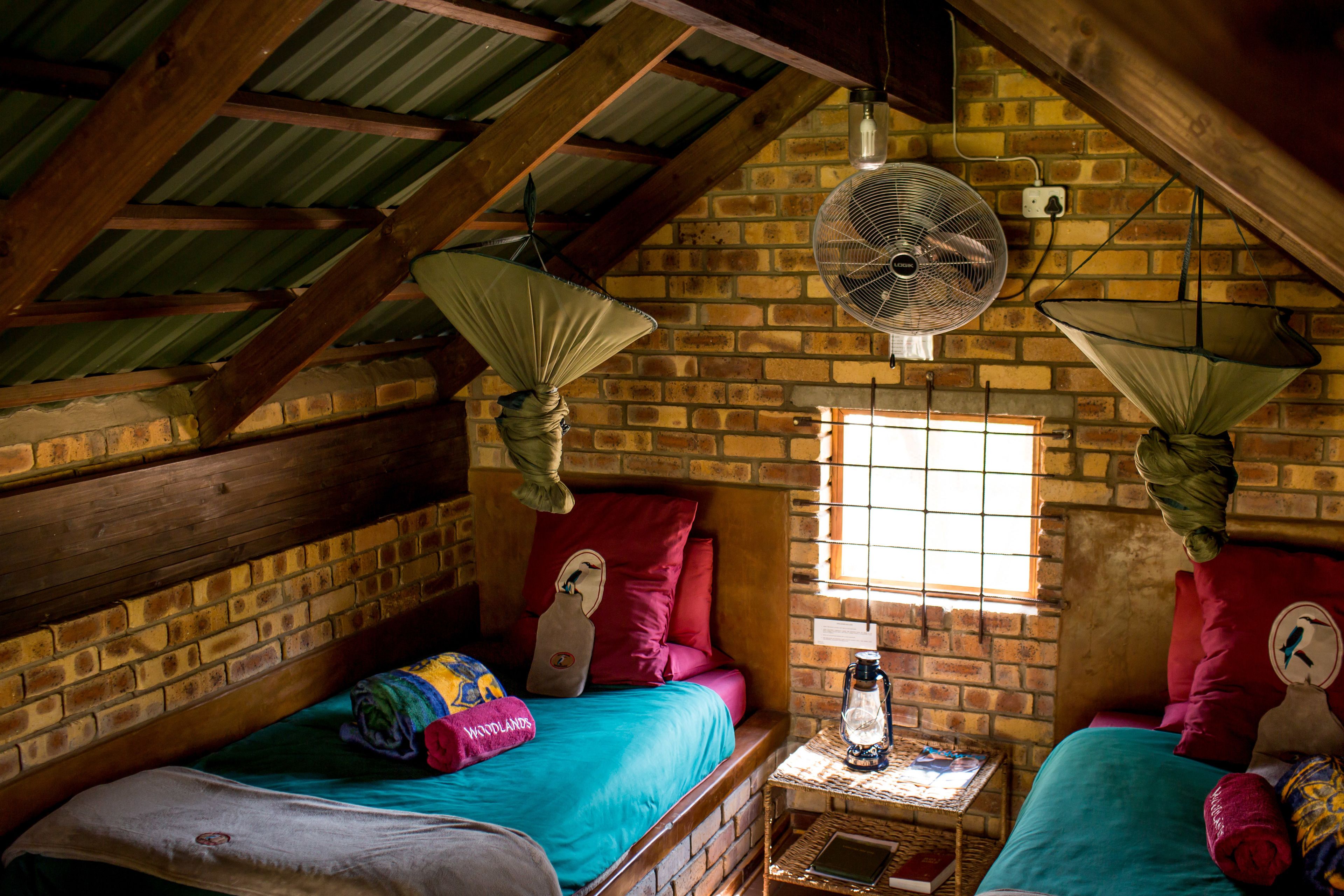 A rustic room with two beds, a brick wall, a wooden ceiling, and a window with a view.