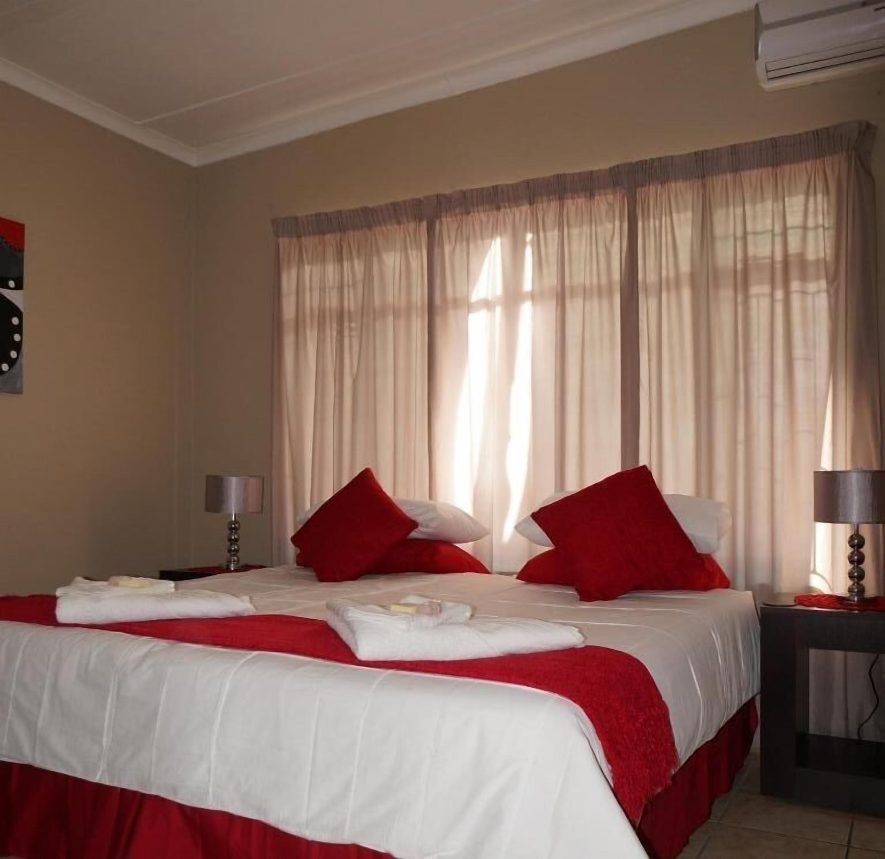 A hotel room with a bed covered in white and red bedding, a window with sheer curtains, and a visible air conditioning unit.
