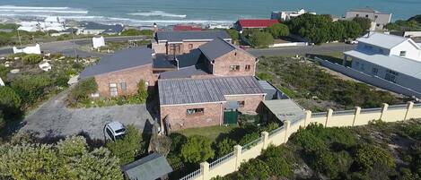 Vista aérea de una propiedad costera con varias edificaciones, incluyendo una estructura de ladrillo destacada con techo inclinado y un edificio más pequeño adyacente con techo plano.