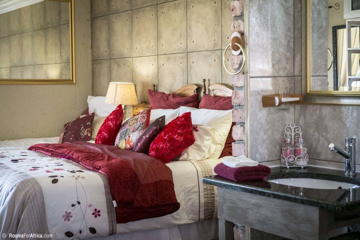 Een slaapkamer met een bed, kussens, een nachtkastje, een lamp, en een badkamer met een wastafel en spiegel.