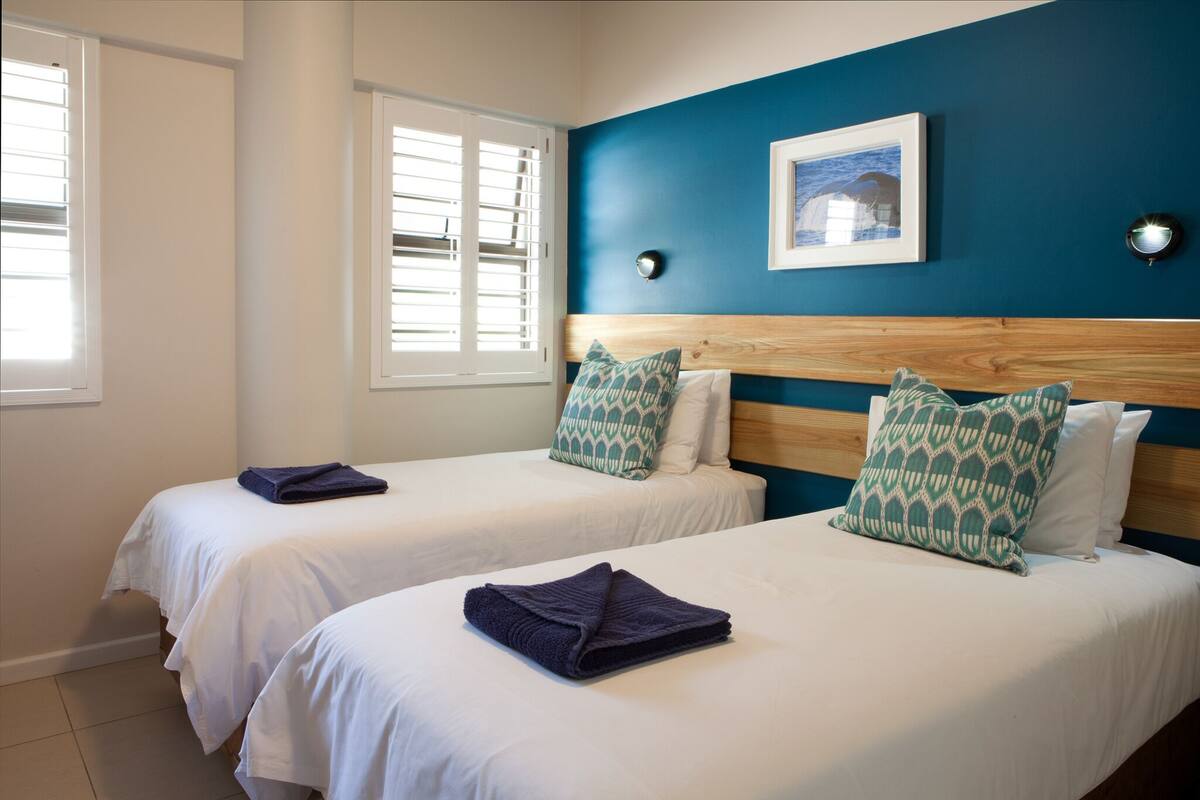 Quarto de hotel com duas camas, roupa de cama branca e paredes azuis.