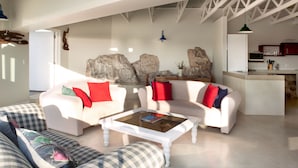 A spacious living area with white sofas, a coffee table, and a kitchen area with red cabinets. - Plett Beachfront Accommodation (Plettenberg Bay)
