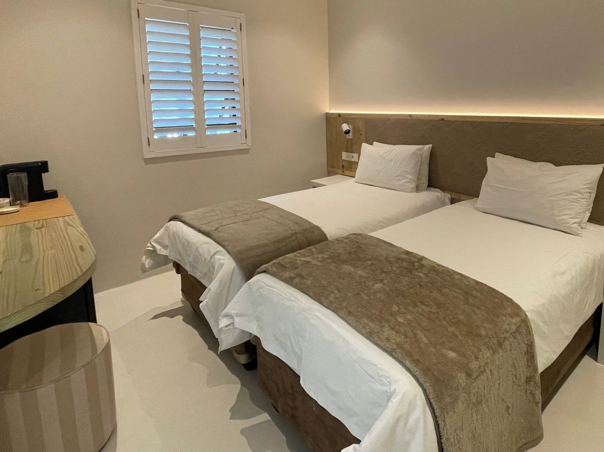 Une chambre d’hôtel avec deux lits, une table de chevet avec une lampe, une poubelle et une fenêtre avec des stores.