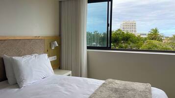 Habitación de hotel con cama, mesita de noche, lámpara y ventana con vista a edificios y árboles.