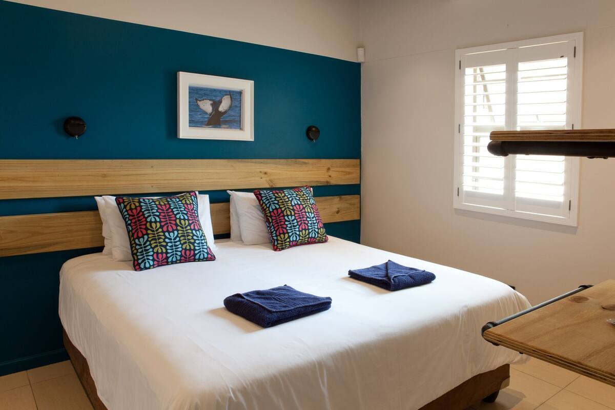 Cama bem arrumada com roupas de cama brancas e azuis, parede de destaque verde-água e quadro acima da cabeceira.