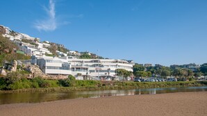A modern hotel with multiple floors, a beach view, and a parking area with cars. - Plett Beachfront Accommodation (Plettenberg Bay)