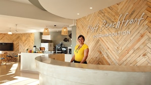A reception area with a herringbone wood wall, a modern kitchen in the background, and a person standing at the reception desk. - Plett Beachfront Accommodation (Plettenberg Bay)
