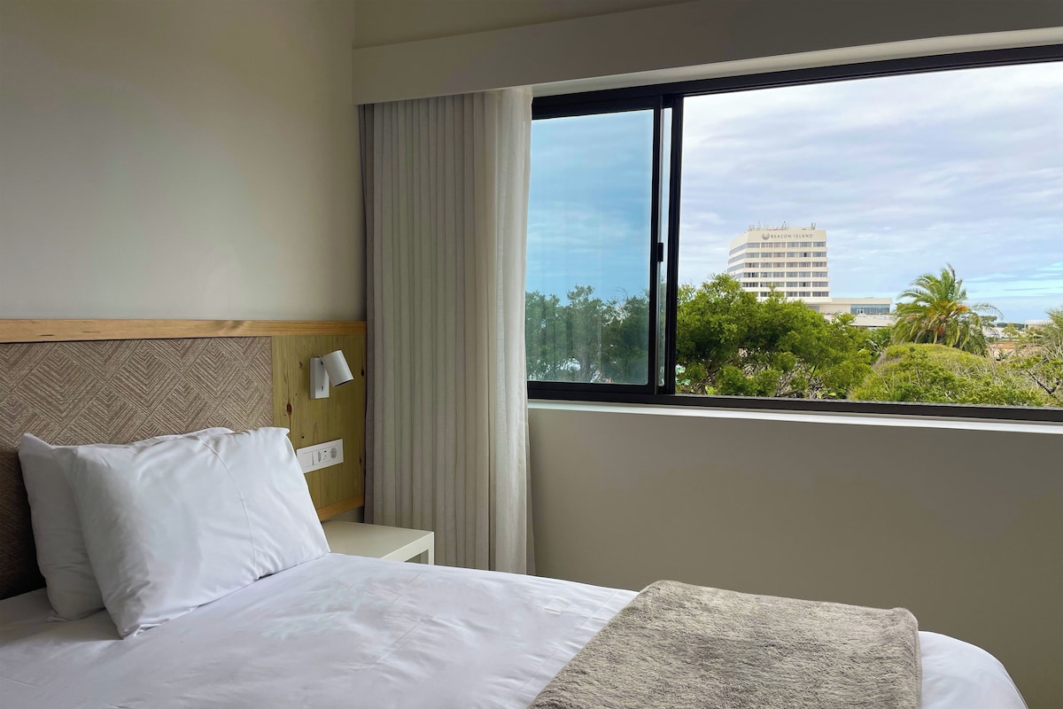 Habitación de hotel con cama, mesita de noche, lámpara y ventana con vista a edificios y árboles.