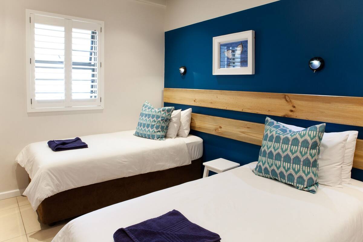 Quarto de hotel com duas camas, cada uma com lençóis brancos e almofadas azuis estampadas. Pequena mesa de cabeceira com abajur em cada lado das camas.
