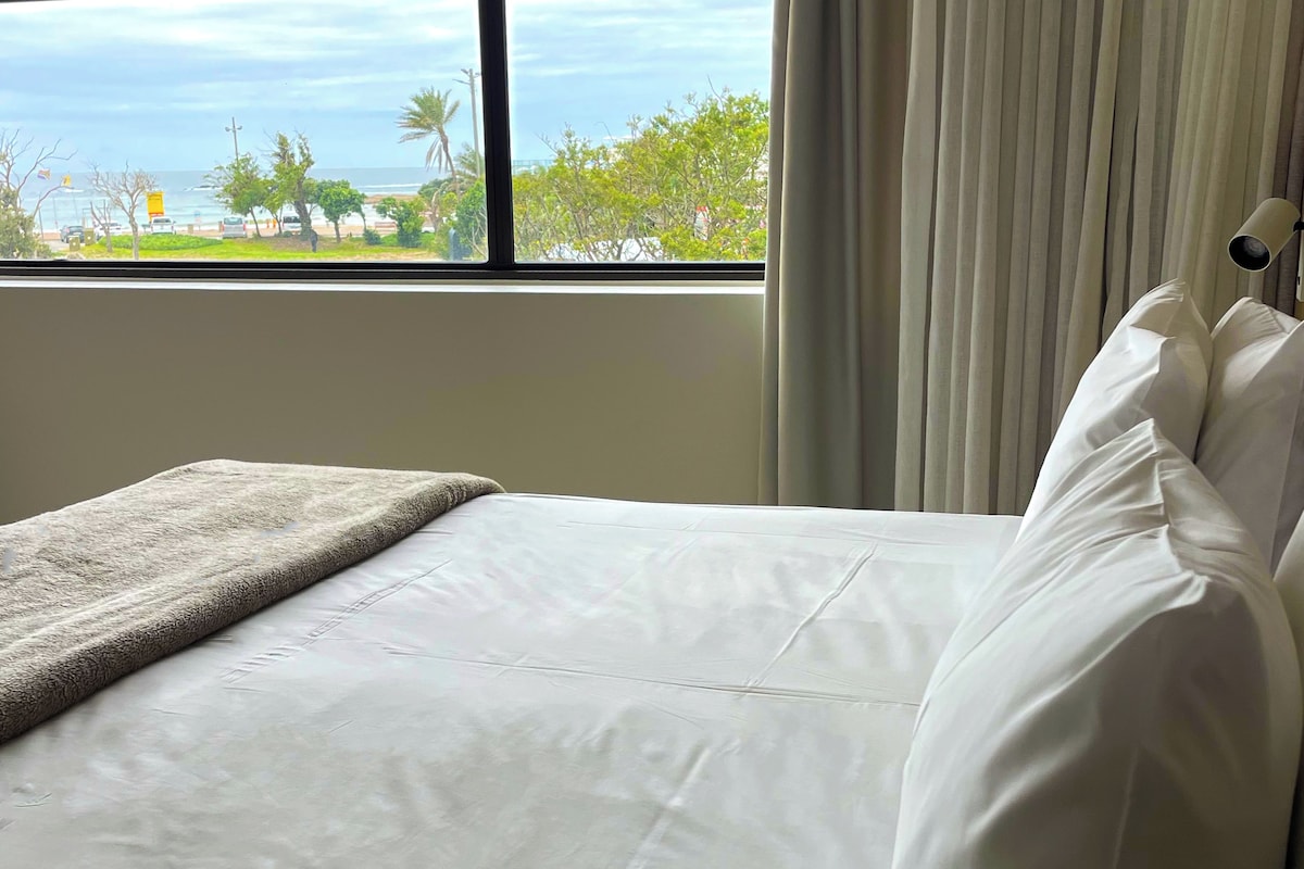 Una cama con ropa de cama blanca y una manta gris, ubicada junto a una ventana con vistas al mar y al follaje.