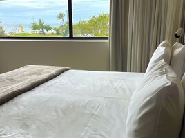 Una cama con ropa de cama blanca y una manta gris, ubicada junto a una ventana con vistas al mar y al follaje.