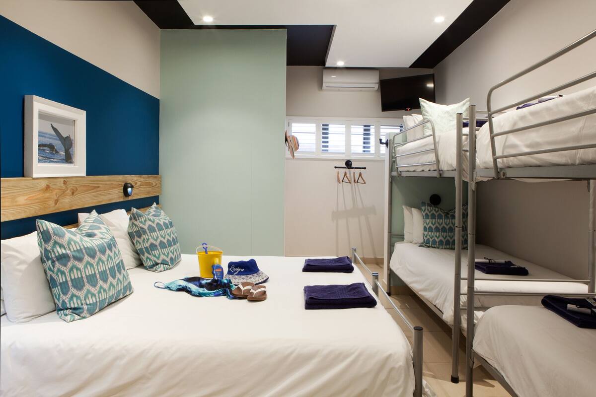 Quarto de hotel com uma cama beliche, uma cama de solteiro e uma cabeceira de madeira. Há um quadro na parede, uma janela e um porta-toalhas.