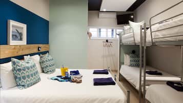 A hotel room with a bunk bed, a single bed, and a wooden headboard. There is a framed picture on the wall, a window, and a towel rack.