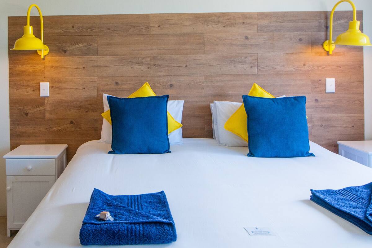 Quarto de hotel com cabeceira de madeira, uma cama grande com roupa de cama branca e duas mesas de cabeceira.