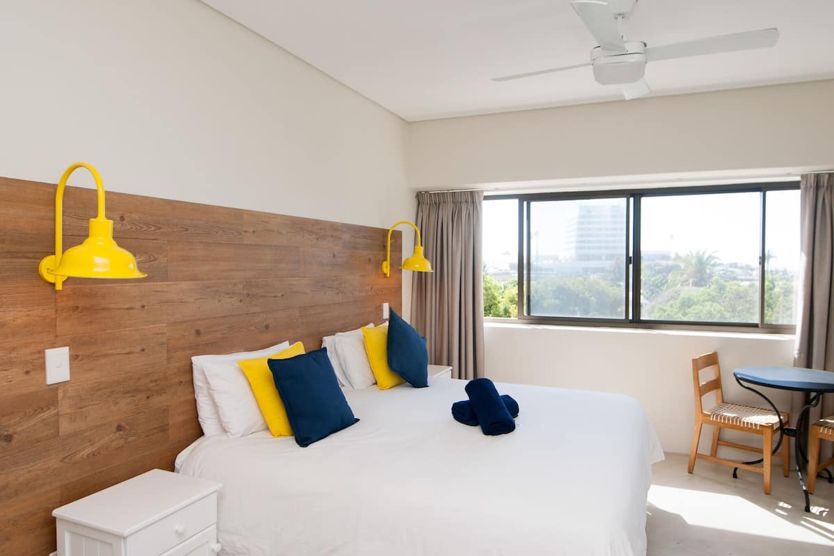 Quarto de hotel moderno com cabeceira de madeira, cama grande com roupa de cama branca e almofadas coloridas, mesa de cabeceira pequena, escrivaninha com cadeira e vista para o verde pela janela.