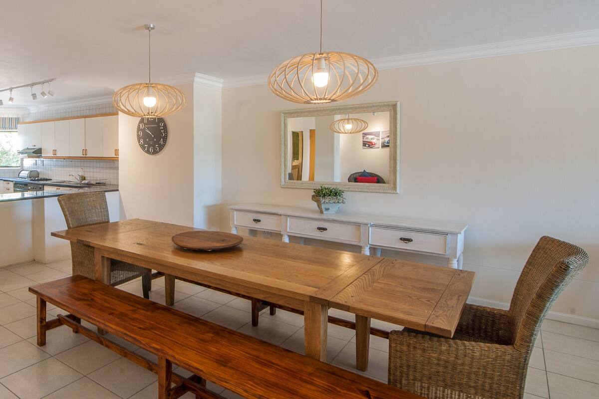 Cozinha moderna com mesa de jantar de madeira, bancos, cadeiras de vime e luminárias pendentes.