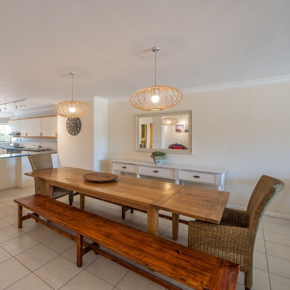 A modern kitchen with a wooden dining table, bench, wicker chairs, and pendant lights.
