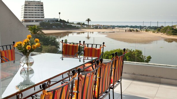 A glass table with striped chairs and a vase of flowers, overlooking a beach and a building labeled "BEACON ISLAND". - Plett Beachfront Accommodation (Plettenberg Bay)