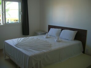 A neatly made bed with white linens and pillows, a bedside table, and a window with curtains.