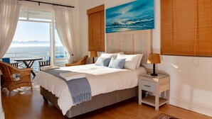 A bedroom with a large bed, a nightstand, a wicker chair, a view of the ocean, and a painting of the ocean. - A Whale of a Time (Cape Town)